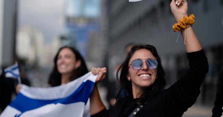 A young woman wearing sunglasses holds an Israeli flag and waves a smaller Israeli flag