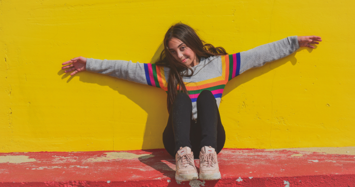 A teen girl against a bright yellow wall with her arms outstretched