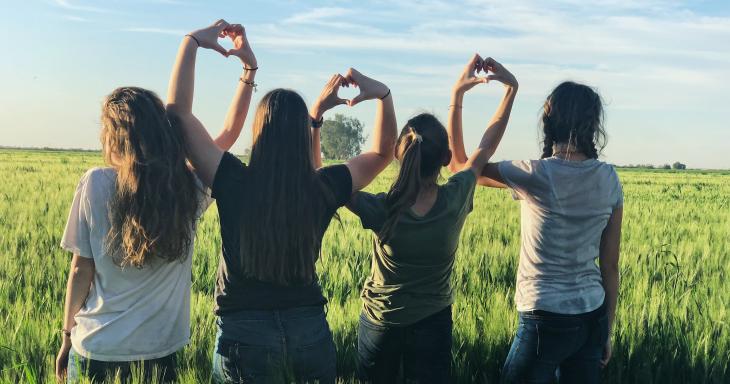 Four children sit in a field with their back to the camera, making hearts with their hands together. 