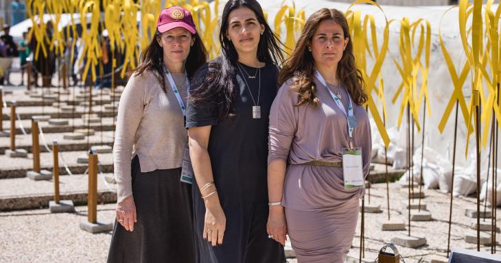 Three women standing together looking solemn with yellow ribbons in the background