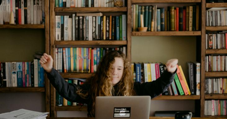 A young girl raises her arms in excitement while using a computer in a library. 
