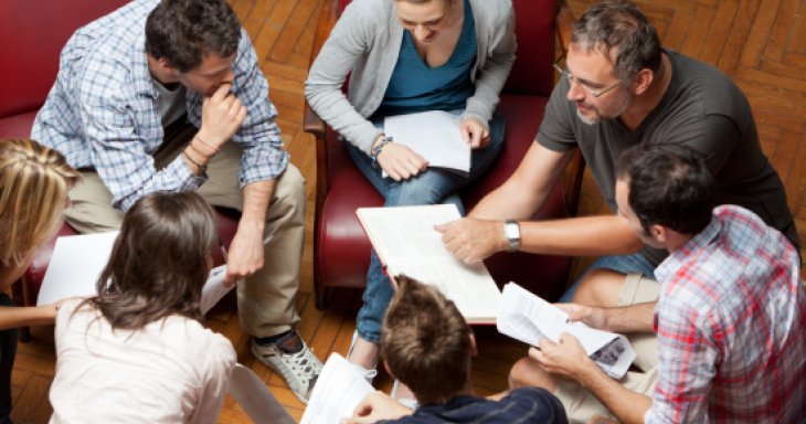 seven teachers - a mix of female-appearing and male-appearing - sit huddled around in a circle with papers