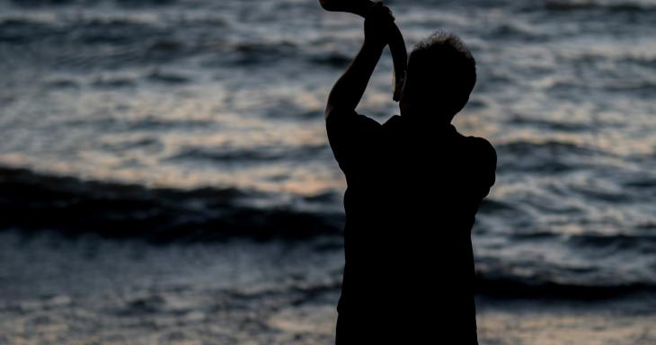 Man blowing shofar at waterfront sunrise