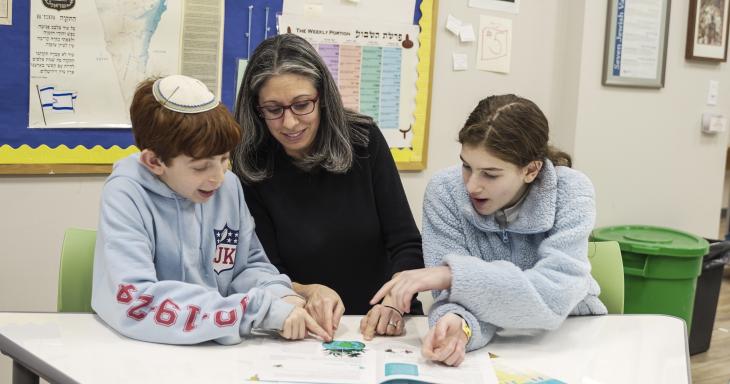 Two children learning with a teacher, a map of the State of Israel in the background.