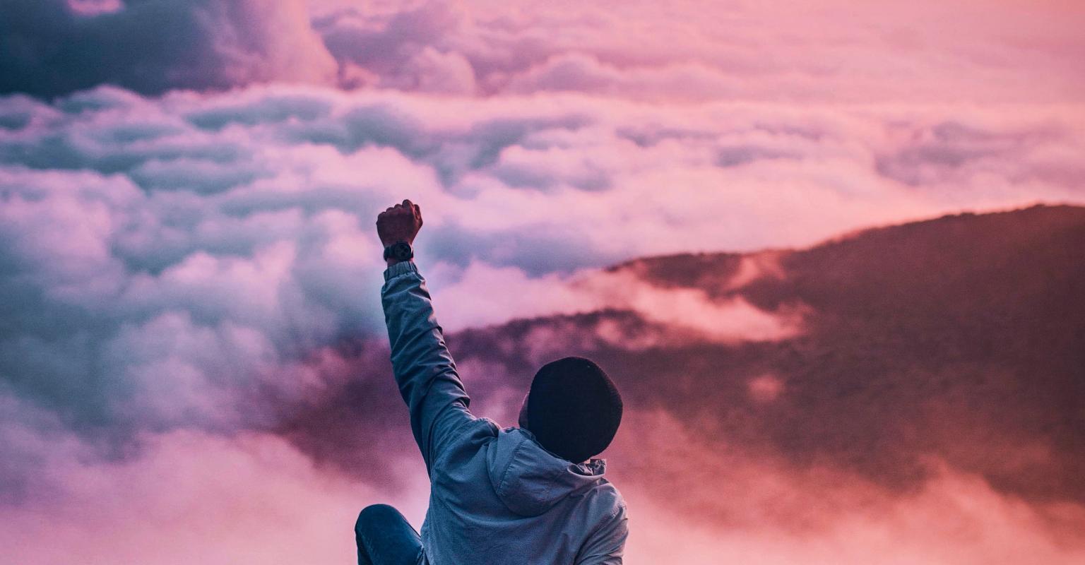 man sitting on mountain cliff facing white clouds rising one hand at golden hour