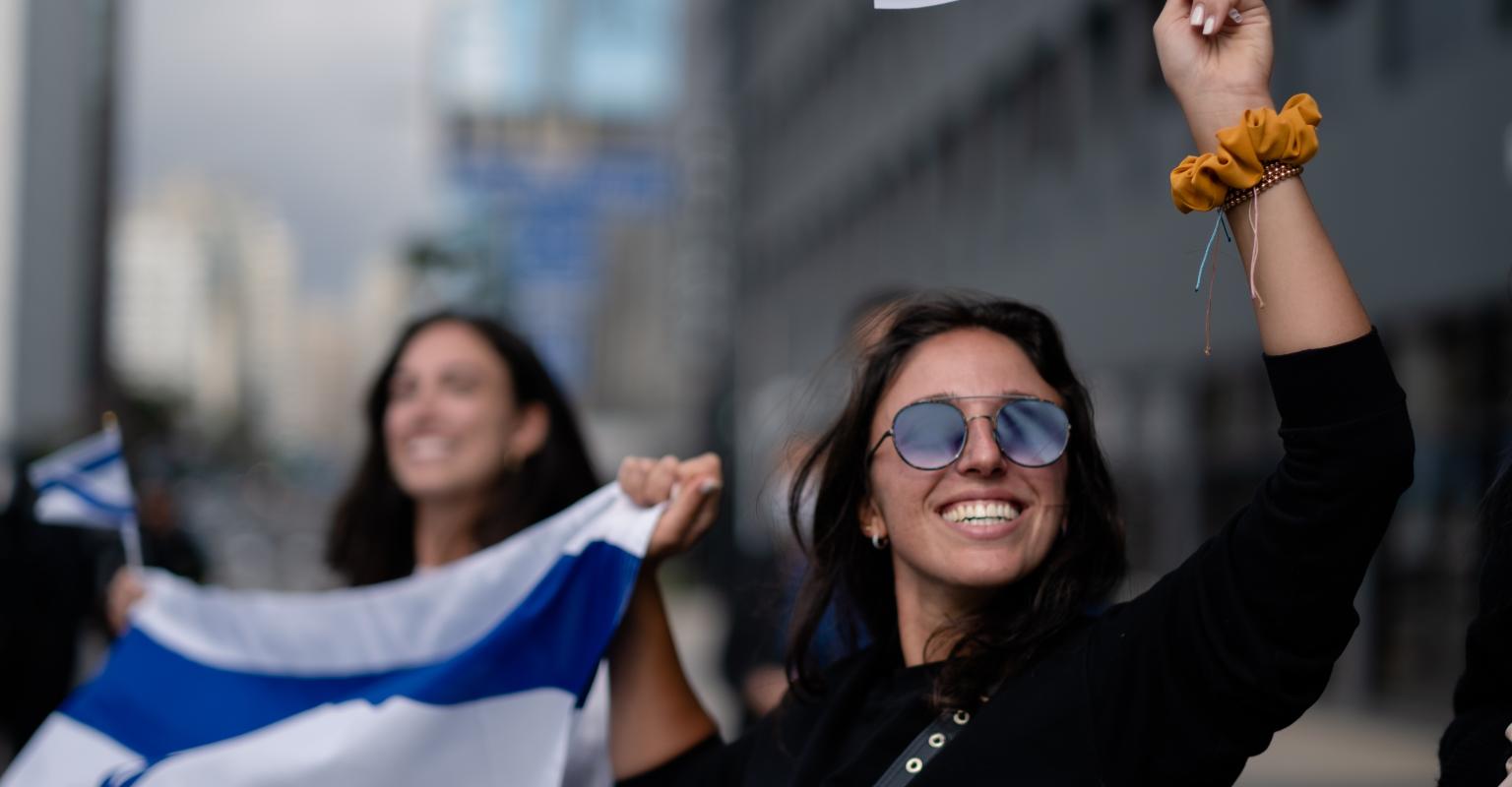 A young woman wearing sunglasses holds an Israeli flag and waves a smaller Israeli flag