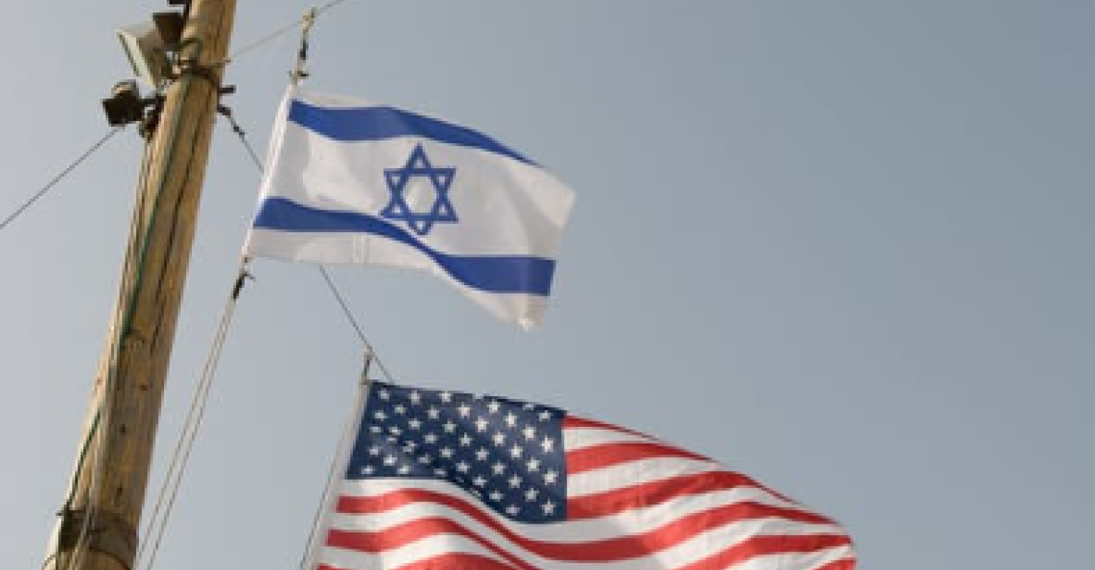 An Israeli flag attached to a pole waves above an American flag