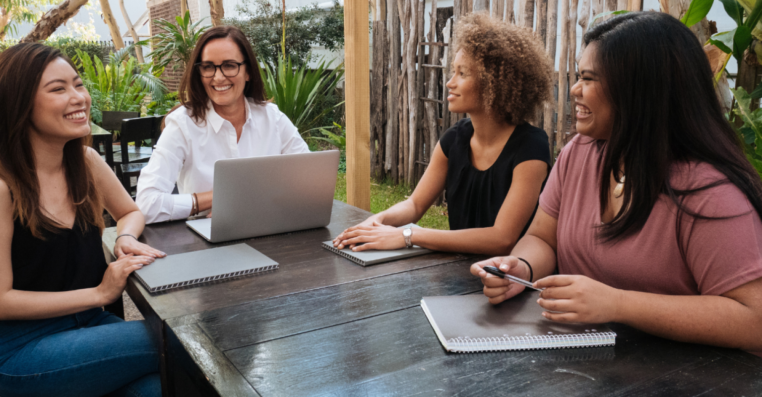 Four women sit at a table outside with notebooks and a laptop
