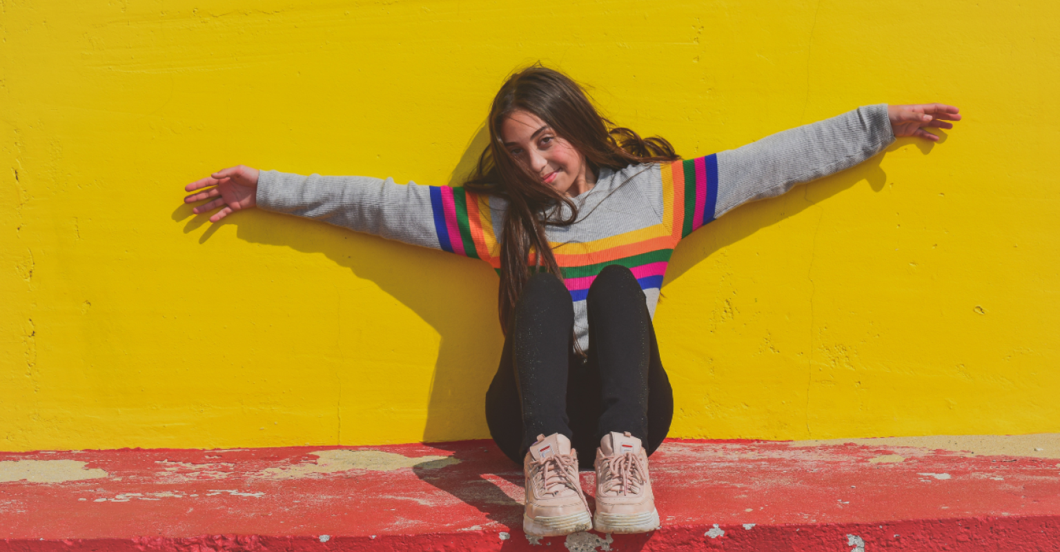 A teen girl against a bright yellow wall with her arms outstretched