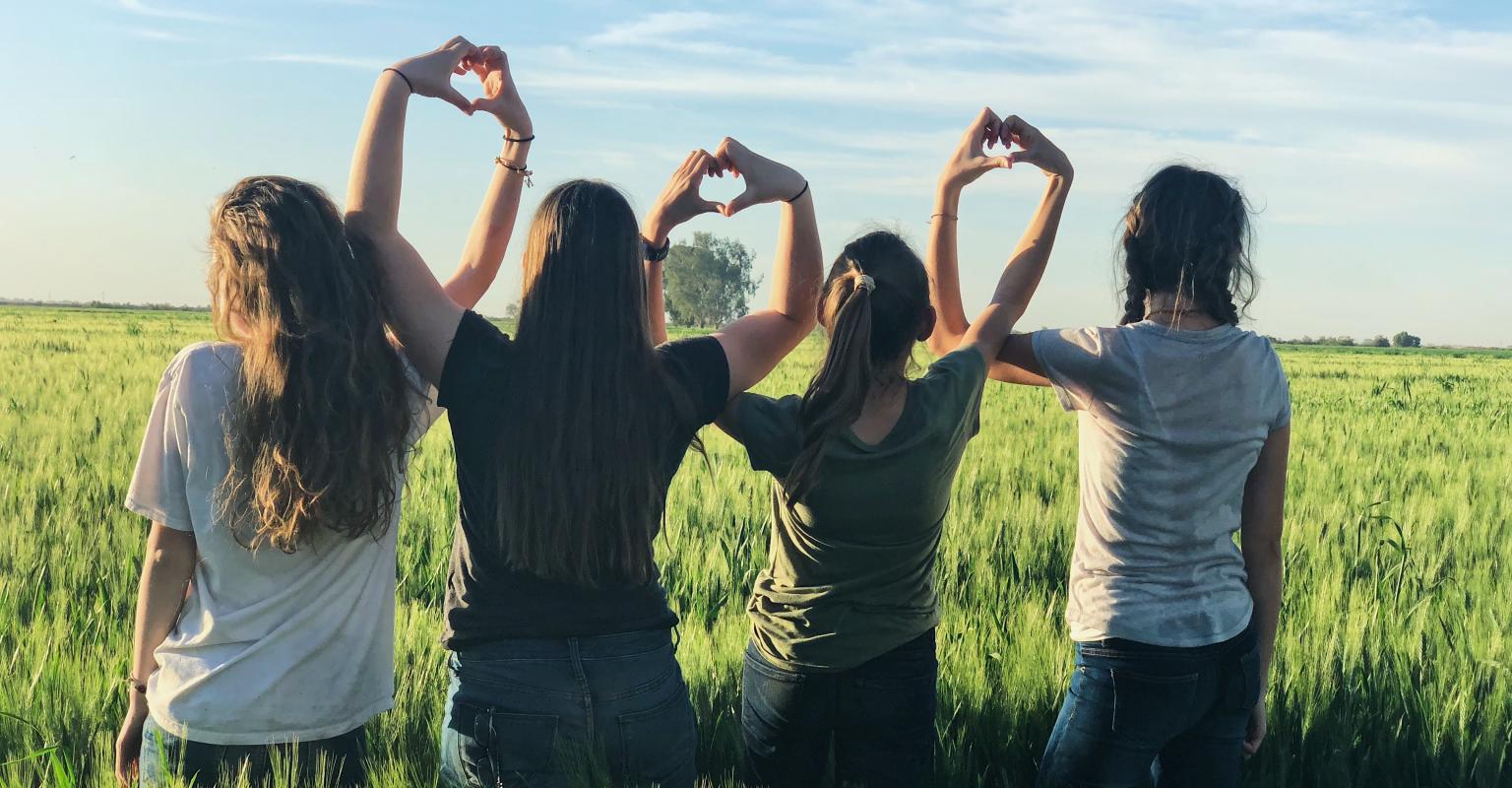 Four children sit in a field with their back to the camera, making hearts with their hands together.