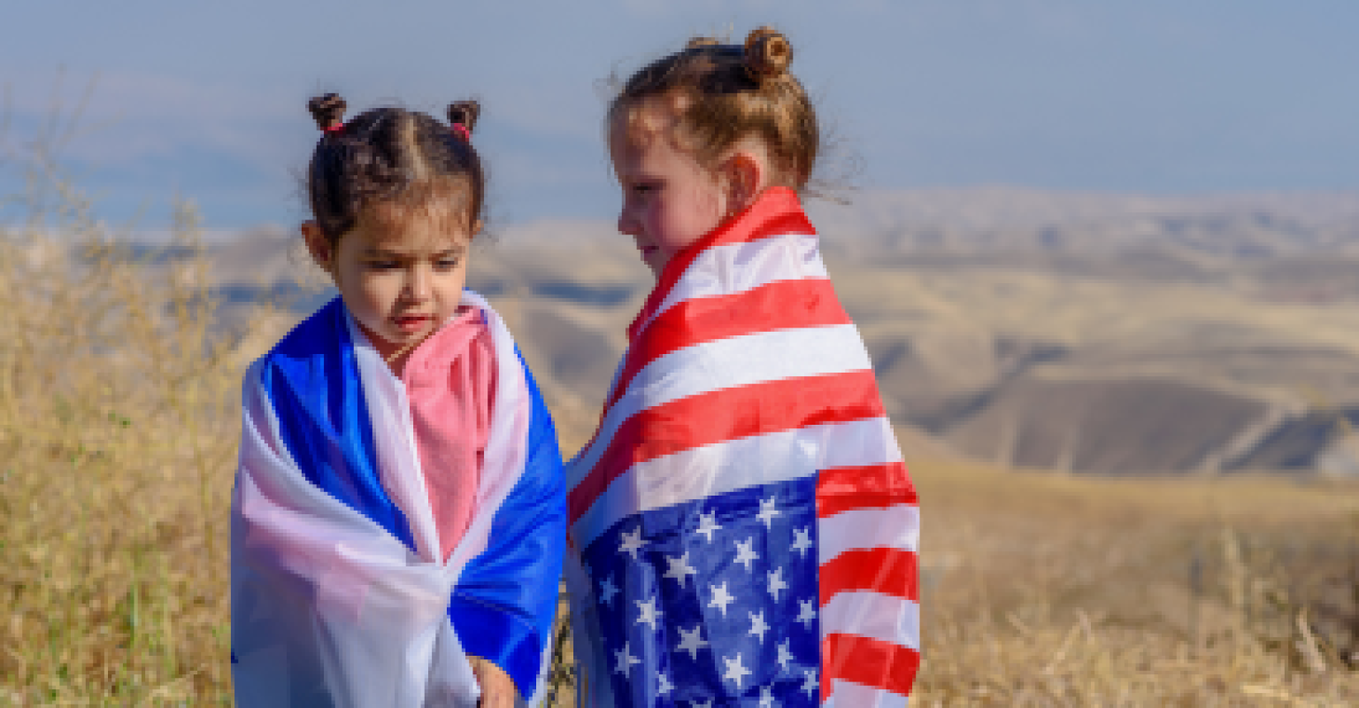 Two young girls standing in a field, one with an Israeli flag wrapped around her and the other with an flag of the US