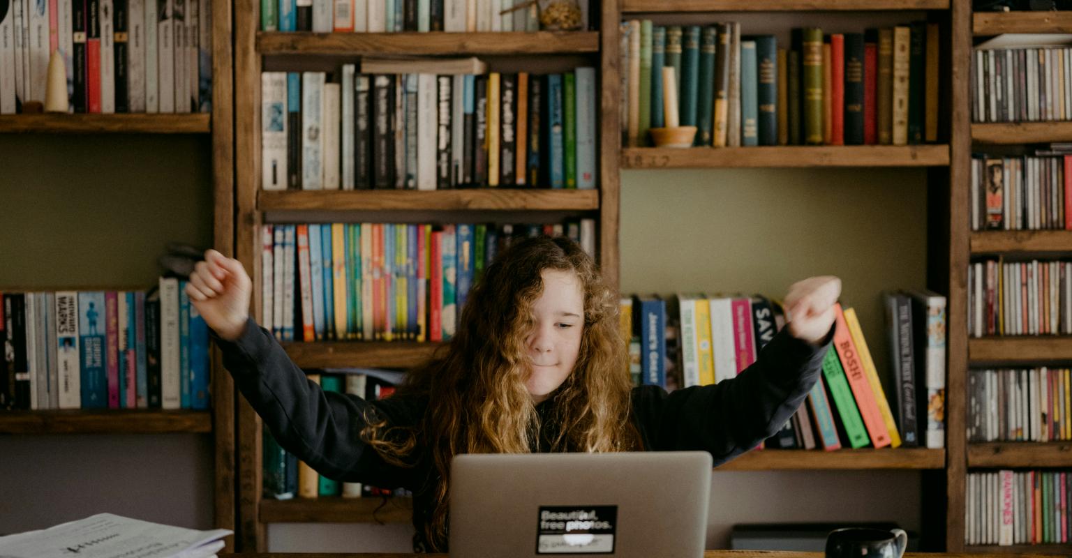 A young girl raises her arms in excitement while using a computer in a library.