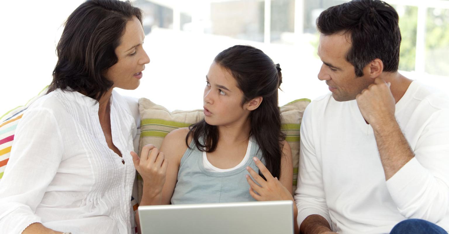 A mother and father look at their teen, who is sitting between them on a couch and talking. They all look concerned.