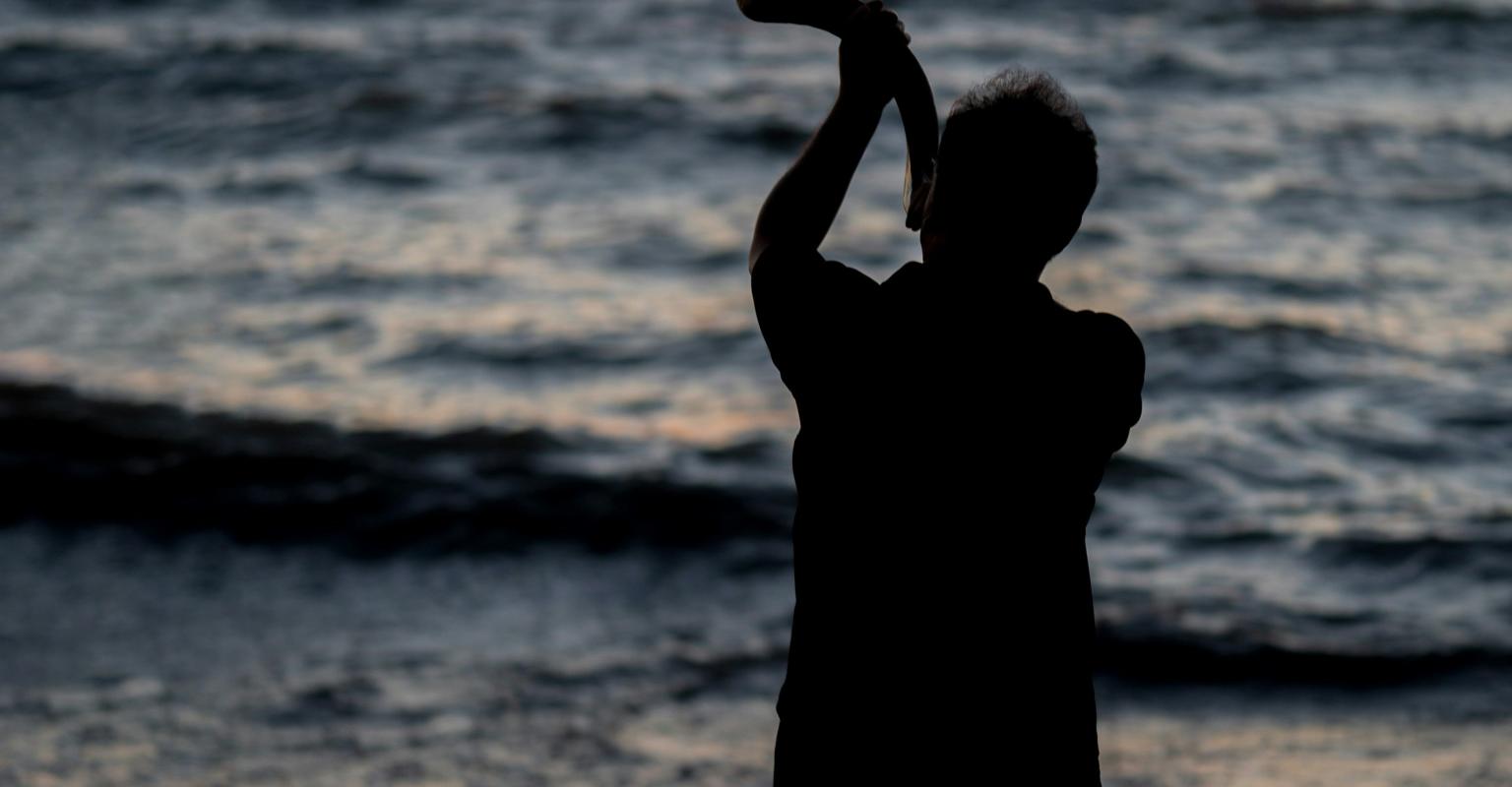 Man blowing shofar at waterfront sunrise