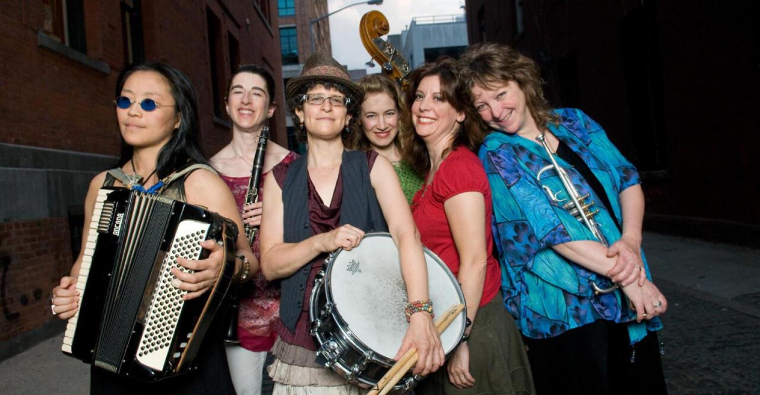 The Isle of Klezbos (L to R): Shoko Nagai, Debra Kreisberg, Photo showing Eve Sicular, Saskia Lane, Melissa Fogarty, and Pam Fleming holding instruments including an accordian, a clarinet, a drum, a trumpet, and a double bass