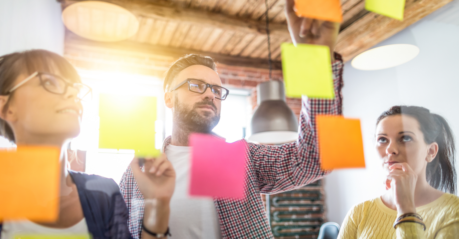 Three excited people examining sticky notes on a glass board.