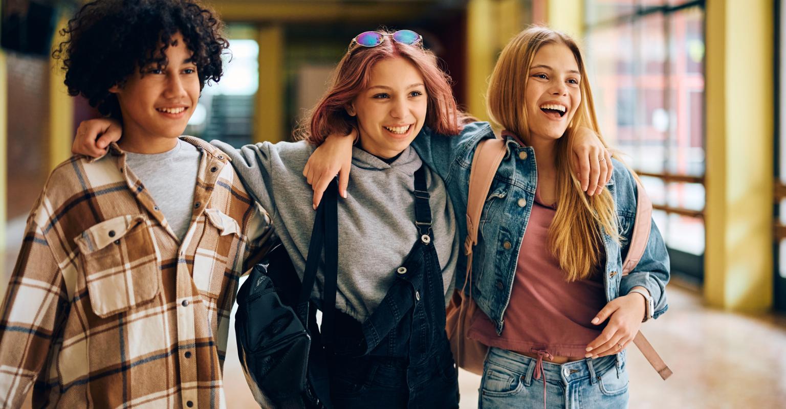 three students - two girls and one boy - walk down a school hallway smiling