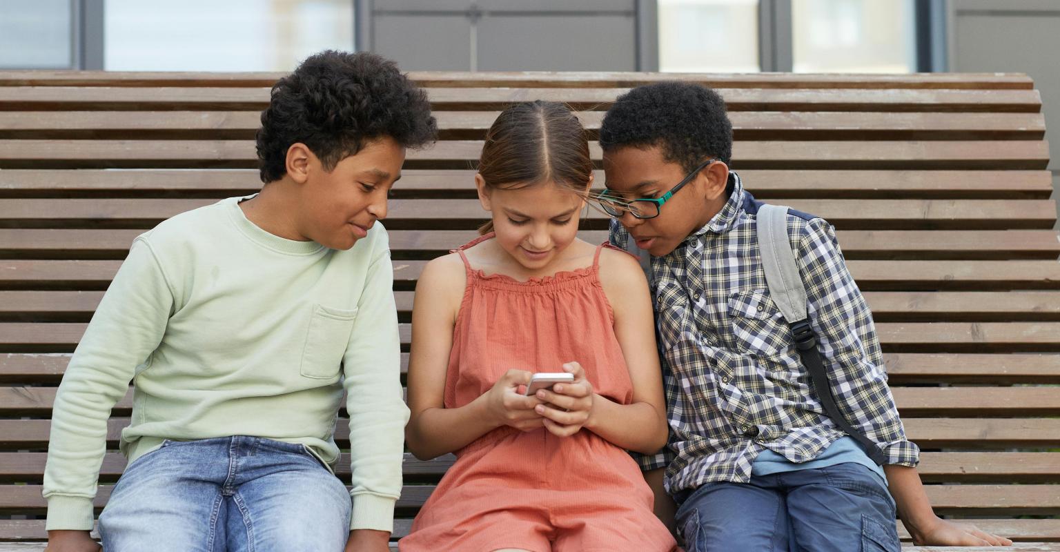 Three children sitting. Child in middle is holding a phone, the others are looking on.