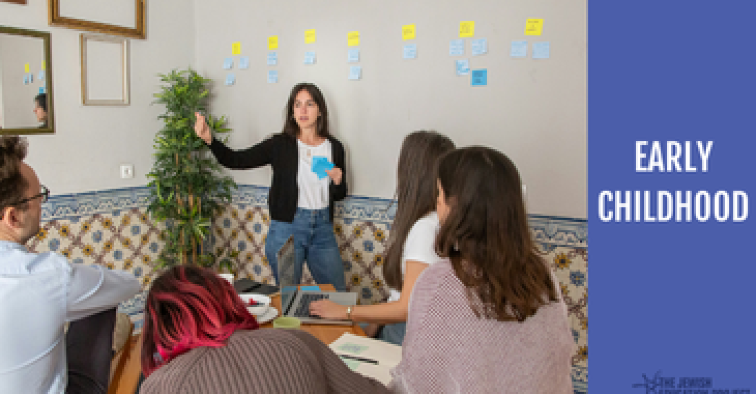 Facilitator standing and speaking with sticky notes behind them. Participants sit at a table. "Early Childhood" banner