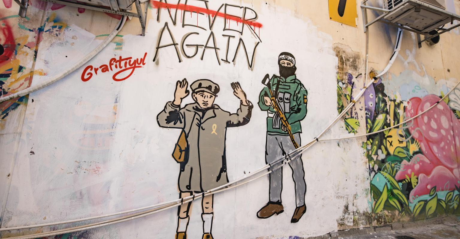 graffiti of hamas militant holding gun next to boy from the holocaust with his hands up, words say "never again" but never is crossed out