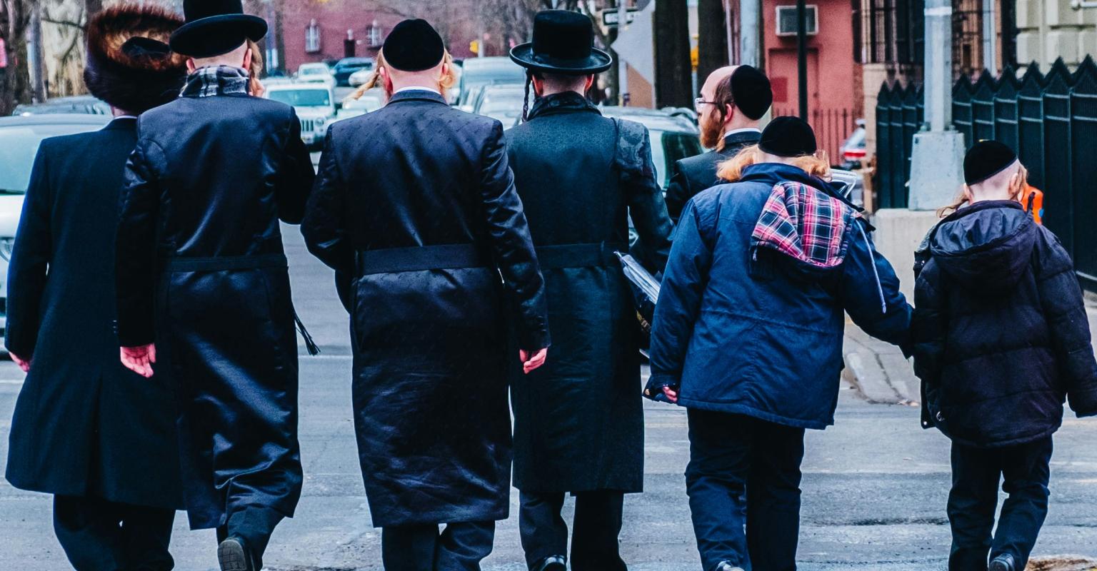 Men and boys in traditional Orthodox Jewish attire walking together through an urban neighborhood.