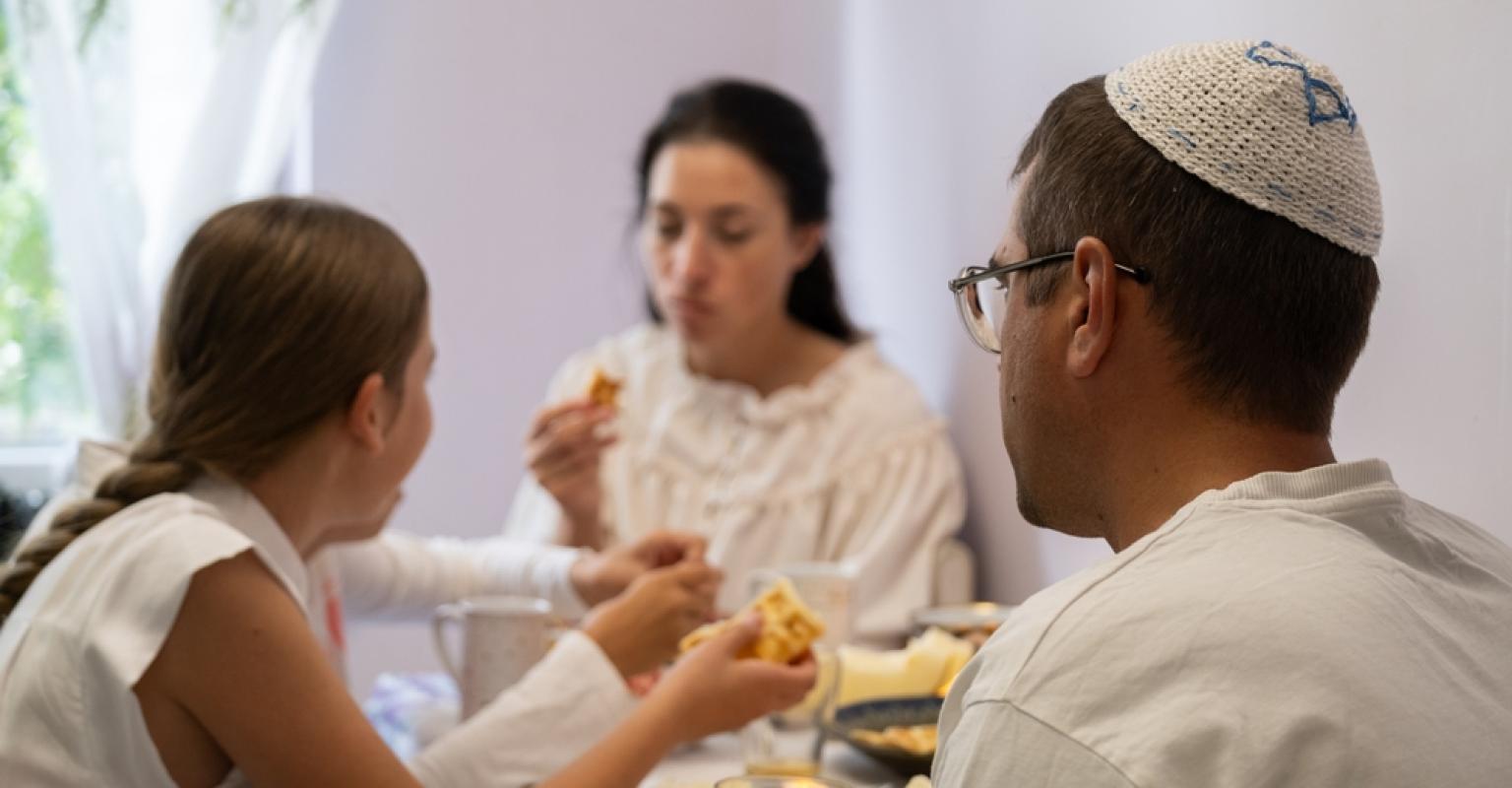 A family sits at a table eating a meal, with a man wearing a kippah and a child sharing food.
