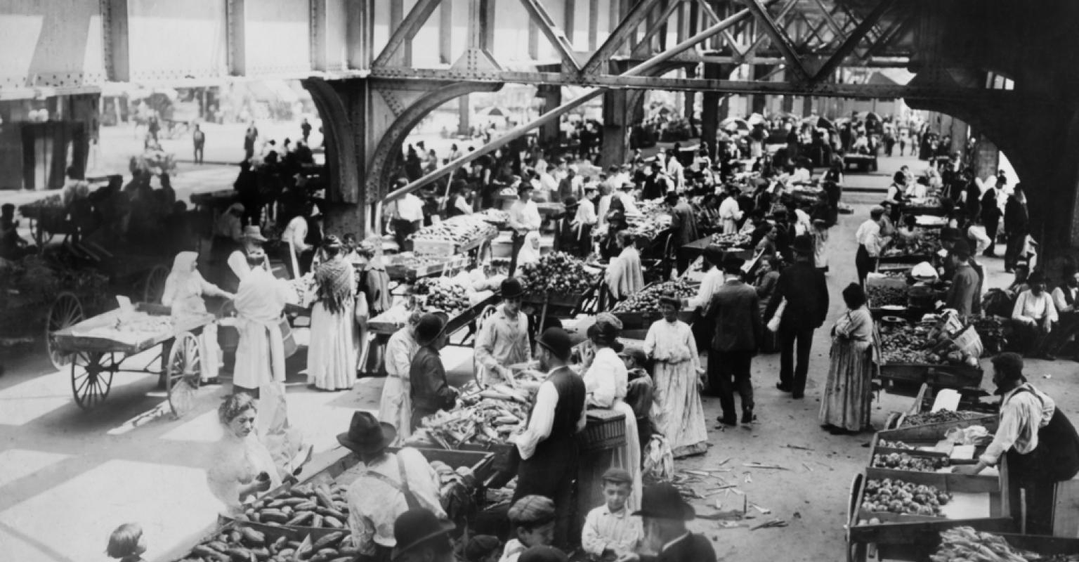 Crowded early 20th-century market under an elevated railway with vendors and shoppers.