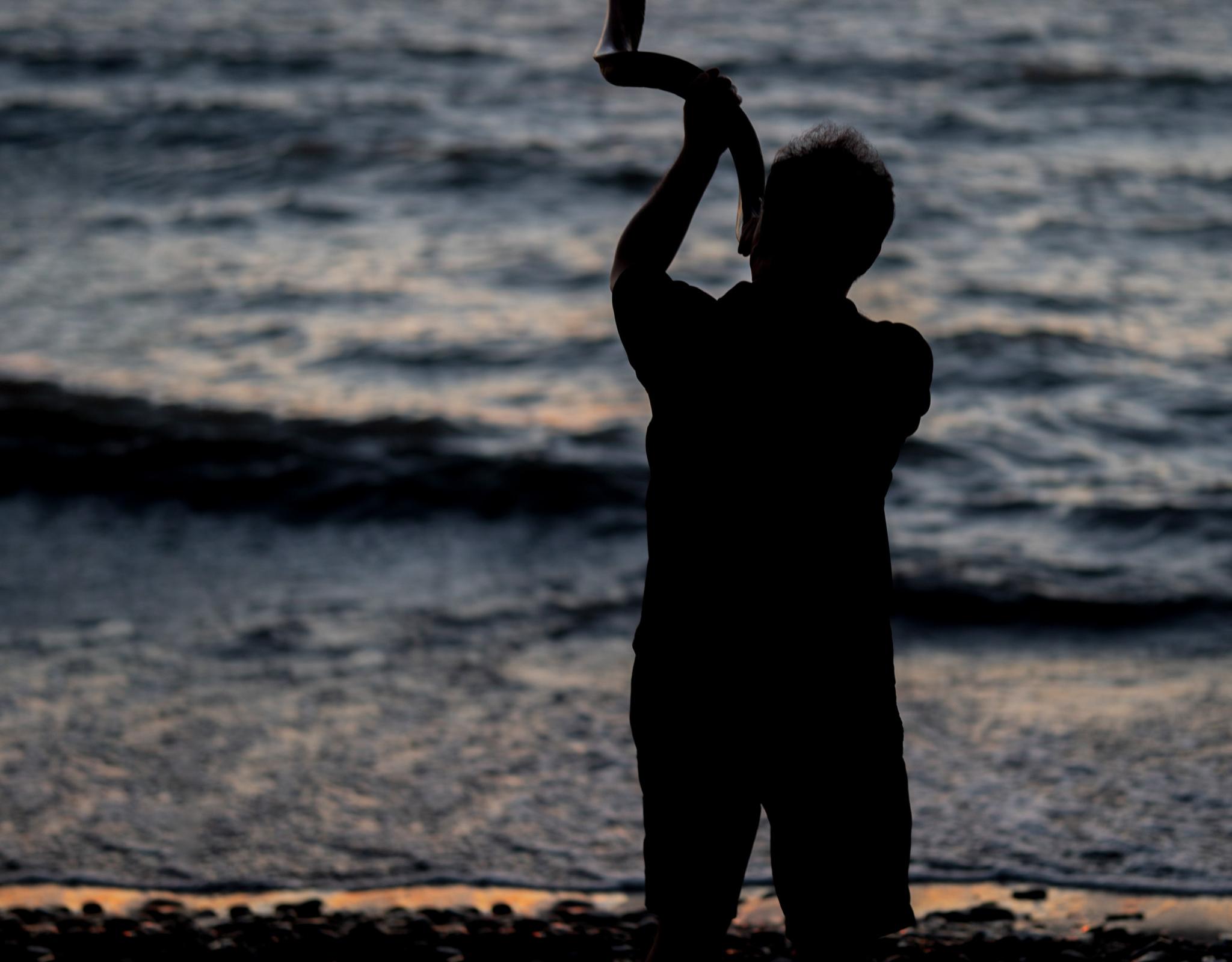 A person blowing the Shofar on a beach at sunset