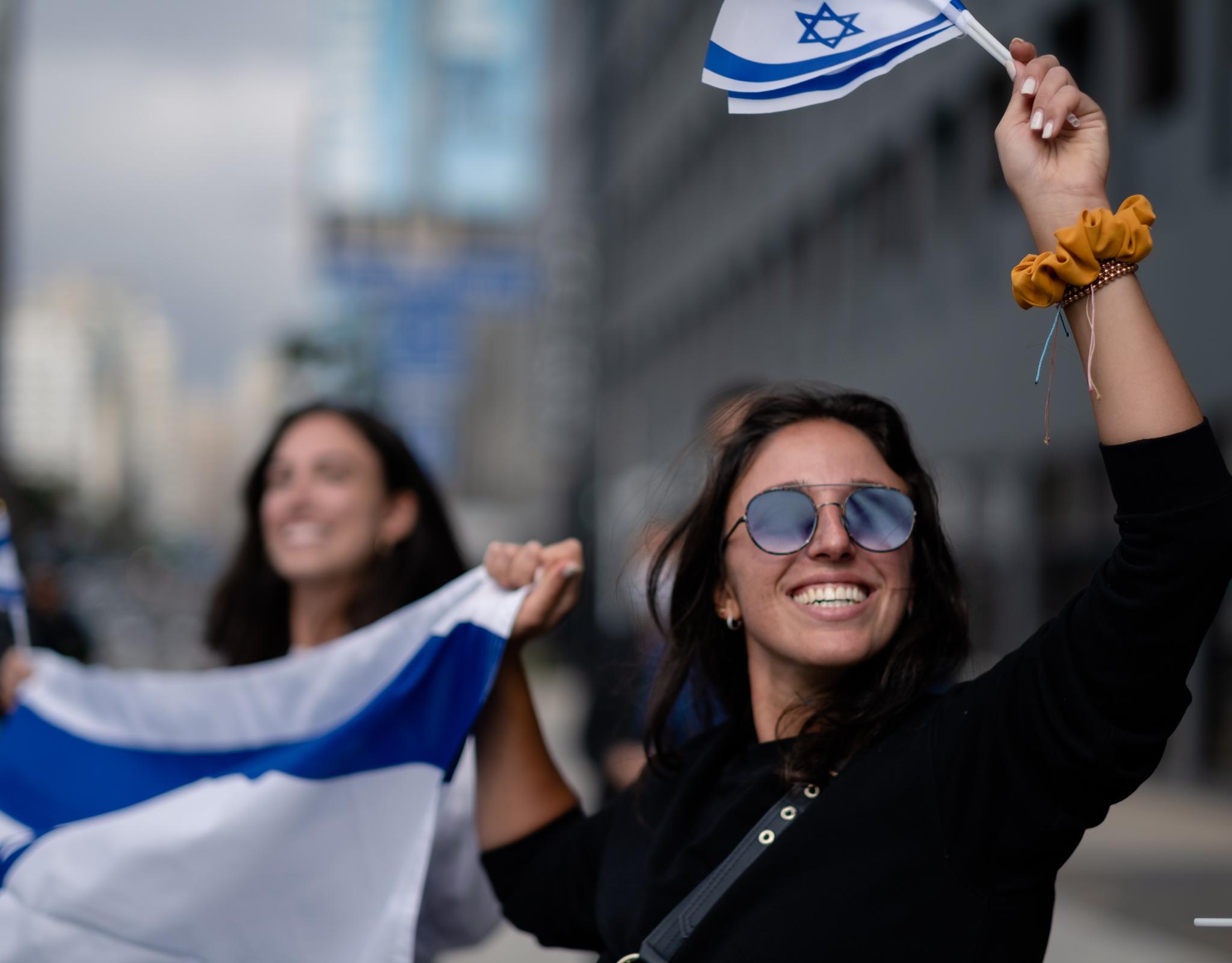 A young woman wearing sunglasses holds an Israeli flag and waves a smaller Israeli flag
