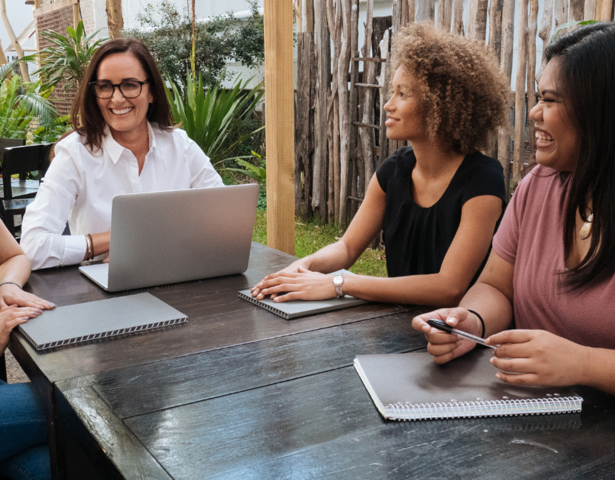 Four women sit at a table outside with notebooks and a laptop