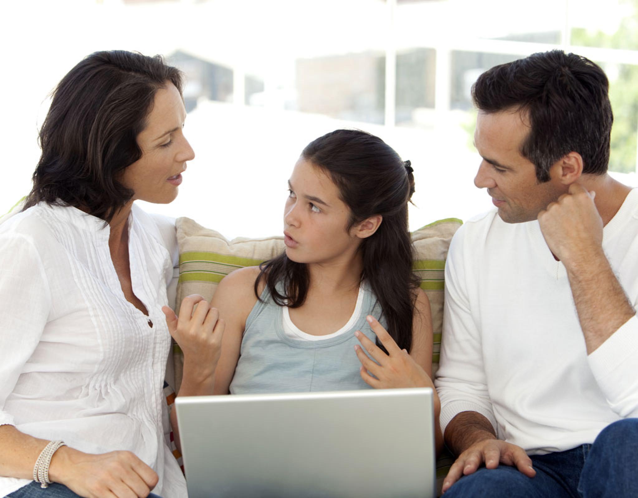 A mother and father look at their teen, who is sitting between them on a couch and talking. They all look concerned.