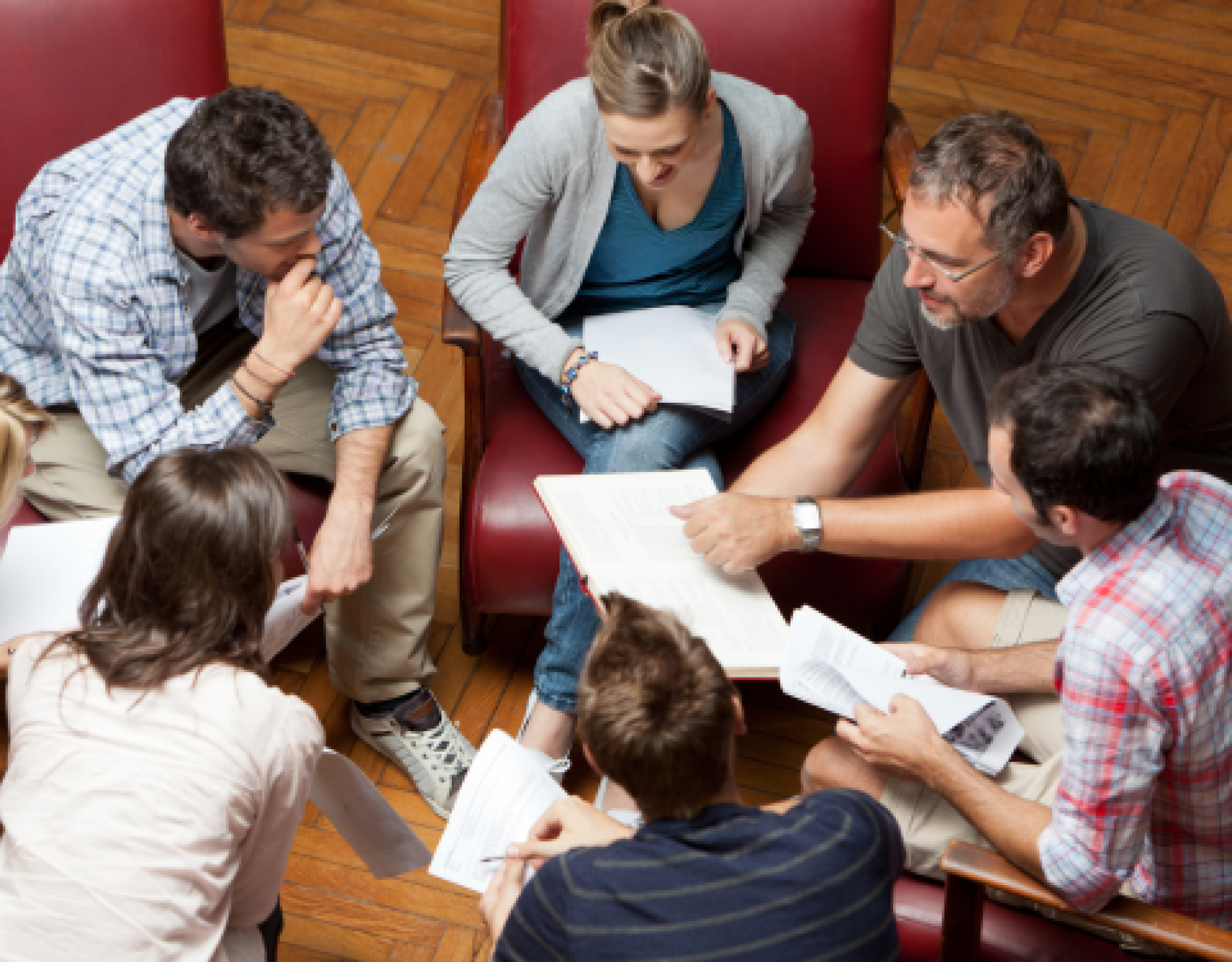 seven teachers - a mix of female-appearing and male-appearing - sit huddled around in a circle with papers