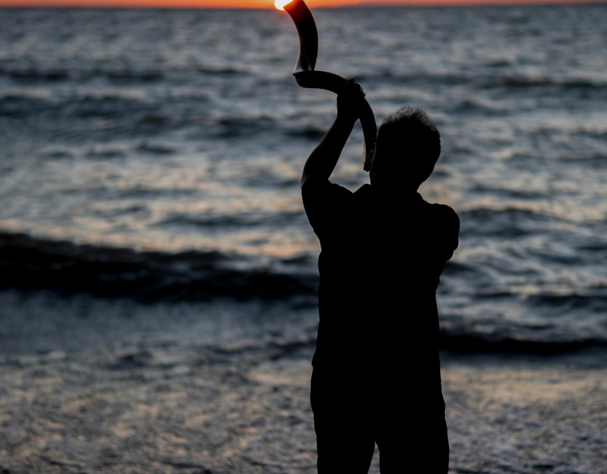 Man blowing shofar at waterfront sunrise
