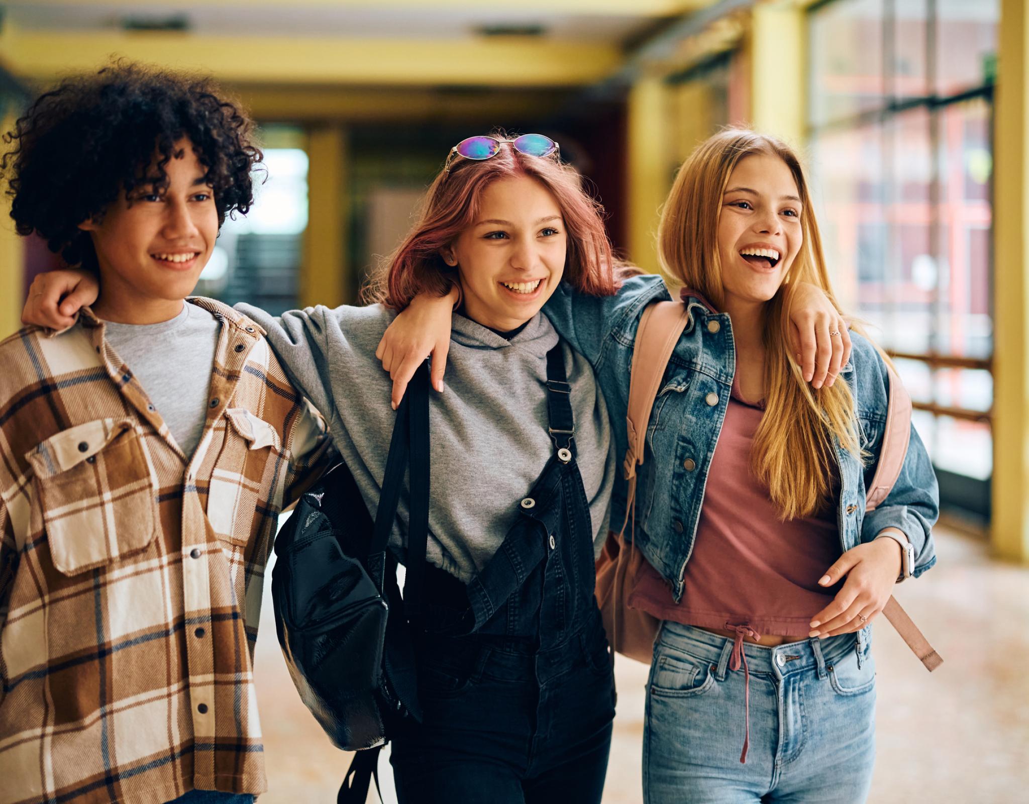 three students - two girls and one boy - walk down a school hallway smiling