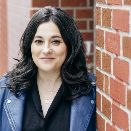 A headshot of Elana leaning against a brick wall.